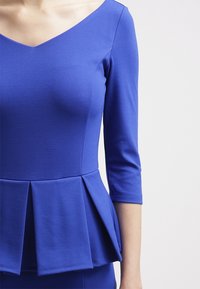 Sininen peplum-toppi, jossa on V-kaula-aukko, kolmenneljäsosa hihat, teksturoitu kangas ja rypytetty yksityiskohta vyötäröllä.