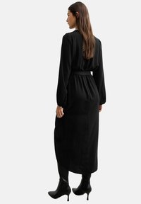 Robe noire à manches longues avec une taille cintrée et une coupe décontractée, présentant une texture lisse et un drapé subtil, accompagnée de bottes à talons noires.