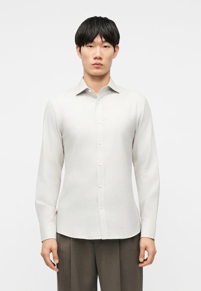 Young man with straight black hair wearing a fitted white button-up shirt and brown pleated trousers, standing against a plain background.