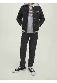 Giacca varsity nera e grigia con strisce bianche, felpa grigia, jeans scuri e sneakers nere con suola bianca. Presenta testo ricamato.