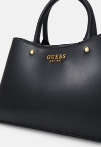 Sac à main en cuir noir avec poignées doubles, clous dorés et logo "GUESS EST 1981" doré à l'avant.