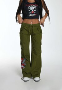 Olijfgroene cargobroek met zijzakken en geborduurde ontwerpen; gecombineerd met een zwarte crop top met een kleurrijke doodskopgrafiek.