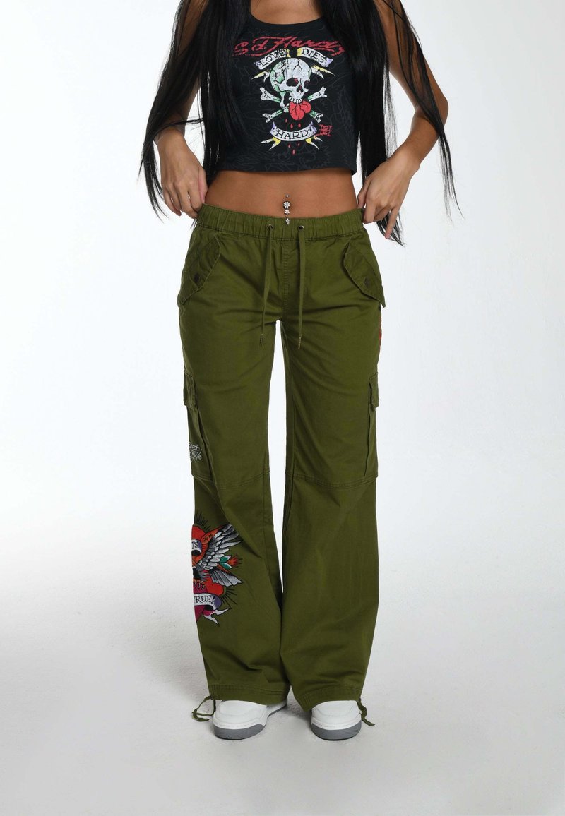 Olijfgroene cargobroek met zijzakken en geborduurde ontwerpen; gecombineerd met een zwarte crop top met een kleurrijke doodskopgrafiek.