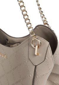 Sac à main gris à surface texturée avec logo embossé. Bandoulière chaîne dorée et accents métalliques. Forme lisse et arrondie avec ouverture supérieure.