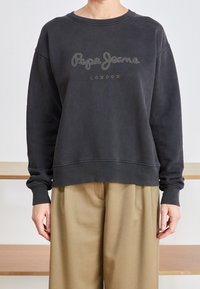 Sudadera negra con cuello redondo, corte holgado y "Pepe Jeans London" en studs decorativos. Combinada con pantalones de pierna ancha beige.