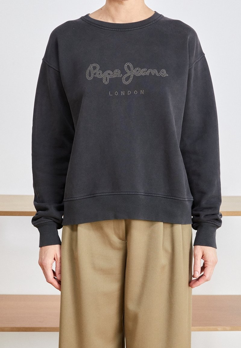 Sudadera negra con cuello redondo, corte holgado y "Pepe Jeans London" en studs decorativos. Combinada con pantalones de pierna ancha beige.