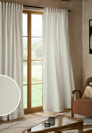 Tende di cotone bianco appese accanto a una porta in vetro con cornice in legno che si affaccia su un'area esterna verde con una poltrona marrone nelle vicinanze.
