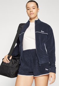 Veste et short de survêtement marine avec des bordures blanches et un logo ; portant un haut rose clair ; tenant un sac en tissu noir avec une sangle.