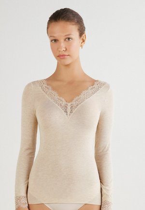 Ung kvinde iført en tætsiddende beige langærmet top med blondedetaljer på V-halsen og manchetterne, stående foran en ensfarvet baggrund.