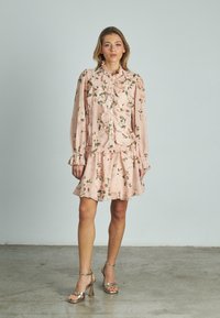 Abito floreale rosa blush con rouches, maniche lunghe e gonna svasata. Abbinato a tacchi metallici con cinturini. Tessuto in satin morbido.