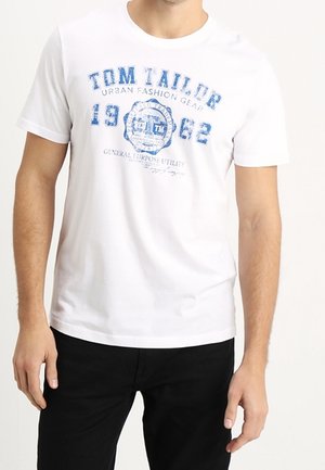 Uomo che indossa una maglietta bianca Tom Tailor con testo e grafiche blu, abbinata a pantaloni neri, su uno sfondo bianco uniforme.