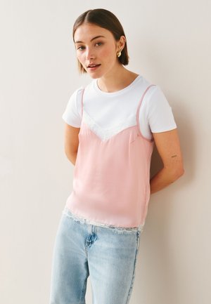 Ung kvinde iført en hvid t-shirt under en lyserød blonder-top og lyseblå jeans, står mod en ensfarvet væg.