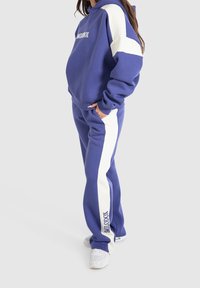 Sudadera con capucha morada con rayas blancas, con logo en el frente. Combina con pantalones deportivos morados con paneles laterales blancos y un ajuste holgado. Zapatillas blancas.