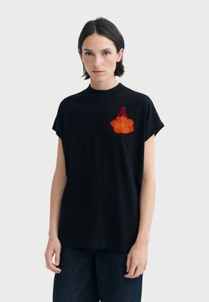 Femme aux cheveux courts et foncés portant un t-shirt noir à manches courtes avec un motif de fruit orange en résille sur le côté gauche de la poitrine, et un pantalon foncé.