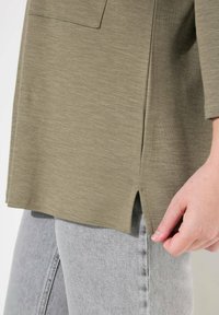 Olivgrünes, langärmeliges Oberteil mit einer Fronttasche und seitlichen Schlitzen. Kombiniert mit hellgrauen, eng anliegenden Jeans. Weicher, strukturierteter Stoff.