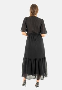 Vestido negro con textura acanalada, mangas cortas y acampanadas, cintura ceñida y dobladillo en capas. Llevado con tacones negros brillantes.