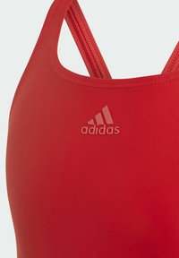 Sujeción deportiva roja con una tela suave, que presenta tirantes finos y un logo de Adidas en relieve en la parte frontal. Sin patrones o acentos adicionales.