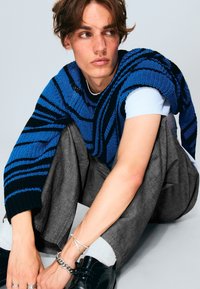 Maglione lavorato a maglia a righe blu e nere indossato sopra una t-shirt leggera, abbinato a pantaloni grigi. Scarpe nere e gioielli in argento.