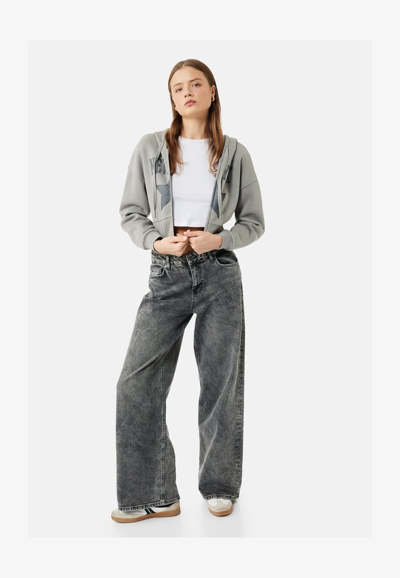 Jeune femme portant un sweat à capuche gris court, un crop top blanc, un jean noir délavé à jambes larges et des baskets blanches, debout les mains jointes.