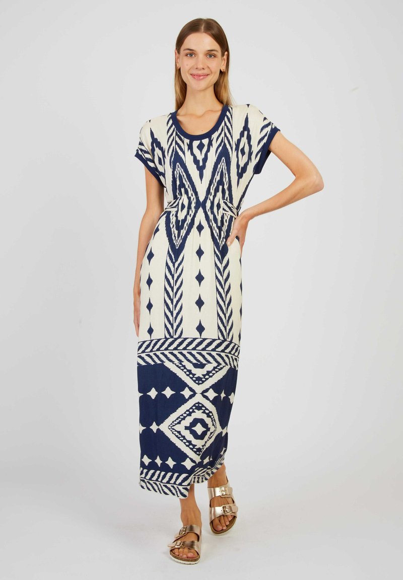 Robe maxi en crème avec des motifs géométriques bleus. Caractéristiques des manches courtes, un col rond et une coupe décontractée. Associée à des sandales métalliques.