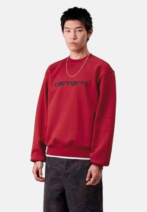 Felpa rossa con polsini e bordo a coste, caratterizzata da logo ricamato in blu scuro. Il modello la indossa con pantaloni scuri a fantasia e una catena argentata.