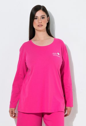 Vrouw met lang donker haar draagt een felroze top met lange mouwen en bijpassende broek, staand tegen een egale lichte achtergrond.