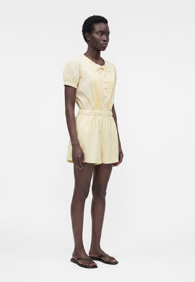 Γυναίκα που στέκεται φορώντας ένα κίτρινο ριγέ ολόσωμο σορτς με κουμπιά και μαύρα επίπεδα σανδάλια, στραμμένη ελαφρώς προς τα αριστερά σε λευκό φόντο.