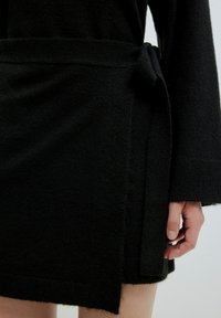Schwarzes Strick-Wickeldress mit strukturiertem Finish, ausgestattet mit einem gebundenen Taillenverschluss und langen Ärmeln, die einen Teil des Arms und des Beins zeigen.