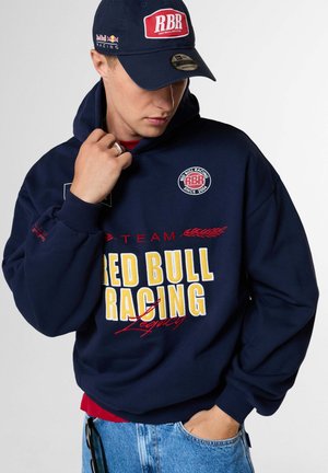 Junger Mann trägt eine marineblaue Red Bull Racing Kappe und Hoodie mit gelb-rotem Logo, blaue Jeans und eine Hand in der Tasche.