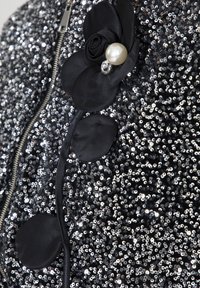 Fiore di tessuto nero con dettagli in perle e gemme su un indumento densamente ricoperto di paillettes argentate accanto a una cerniera metallica.