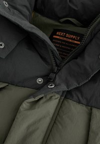 Giacca puffer con un estratto nero e una fodera interna verde oliva, dotata di una cerniera frontale, bottoni a pressione e un'etichetta di marca all'interno.