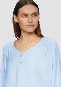 Blouse bleu clair avec un col en V, présentant une texture plissée aux épaules et un détail de bouton. Tissu doux et léger.