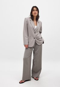 Gaišpelēks blazeris ar pārspīlētu piegriezumu, ar vienu pogu aizdari un divām priekšējām kabatām, savienots ar platām pelēkām biksēm un melniem sandaliem.