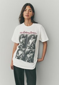 Hvid bomuldstrøje med et sort-hvidt grafik af musikere, med teksten "the Rolling Stones" i maroon over billederne.