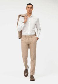 Weißes Hemd mit Knopfleiste, beige Leinenhosen und braunen Mokassins. Das Modell hält eine beige Jacke und präsentiert eine moderne, taillierte Passform.