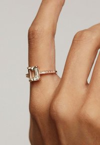 Anillo de oro rosa con una piedra en corte esmeralda de color claro, rodeada de piedras más pequeñas y claras en una delicada banda.