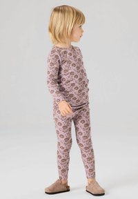 Jeune enfant debout de profil portant un haut à manches longues et un legging assortis à motifs floraux violets, avec des chaussures beiges sans lacets, sur fond gris clair.