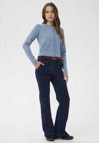 Gaiši zils pogu krekls no mīksta zeķes materiāla, apvienots ar tumši ziliem platām kājām džinsiem ar sānu šuvēm un brūnu jostu.