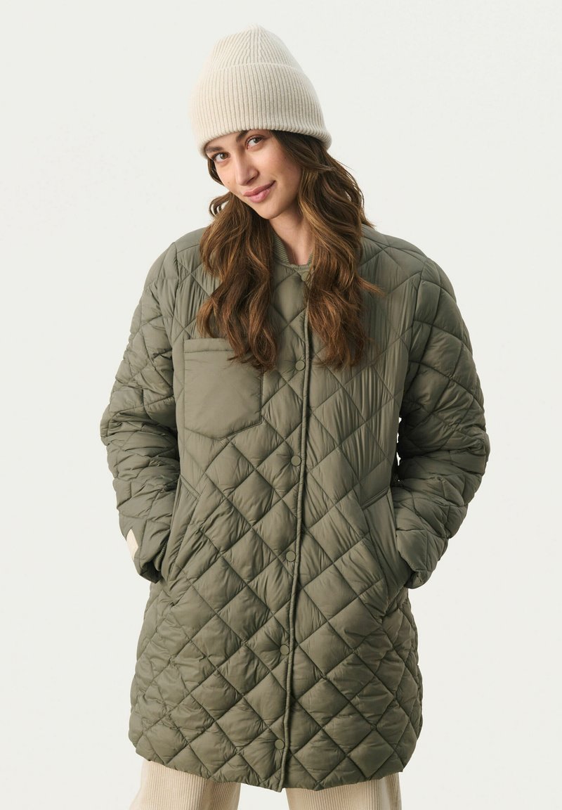 Zielona oliwkowa pikowana długa kurtka z diamentowym wzorem, zapinana na guziki, z bocznymi kieszeniami, w połączeniu z kremową czapką typu beanie z prążkowanego dzianiny.