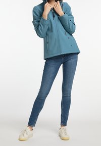 Veste softshell bleu sarcelle avec capuche, dotée d'une fermeture éclair à l'avant et de boutons-pression. Portée avec un jean bleu slim et des baskets blanches.