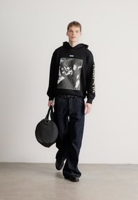 Μαύρο φούτερ με κουκούλα και γραφικό σχέδιο, σκούρο μπλε τζιν και μαύρα παπούτσια. Κρατάει μια στρογγυλή μαύρη τσάντα. Απλό, καθημερινό στυλ δρόμου.