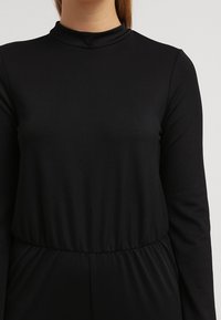 Top negro de manga larga con cuello alto, hecho de tejido suave. El diseño presenta una cintura ajustada y una textura suave. No hay patrones ni elementos metálicos visibles.