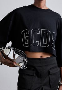 Camiseta corta negra con logo "GCDS" de strass, combinada con un bolso de mano negro y plateado brillante con correa de cadena.