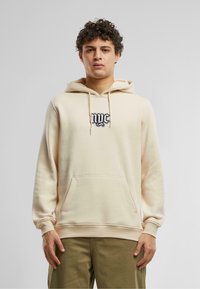 Felpa beige con tasca a marsupio e cappuccio con cordino. Presenta una grafica nera di NYC ricamata nella zona del petto. Tessuto morbido.