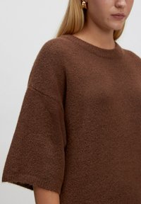 Brauner Strickpullover mit rundem Ausschnitt und kurzen, weiten Ärmeln. Die Textur ist weich und leicht flauschig, was ein gemütliches Aussehen verleiht.