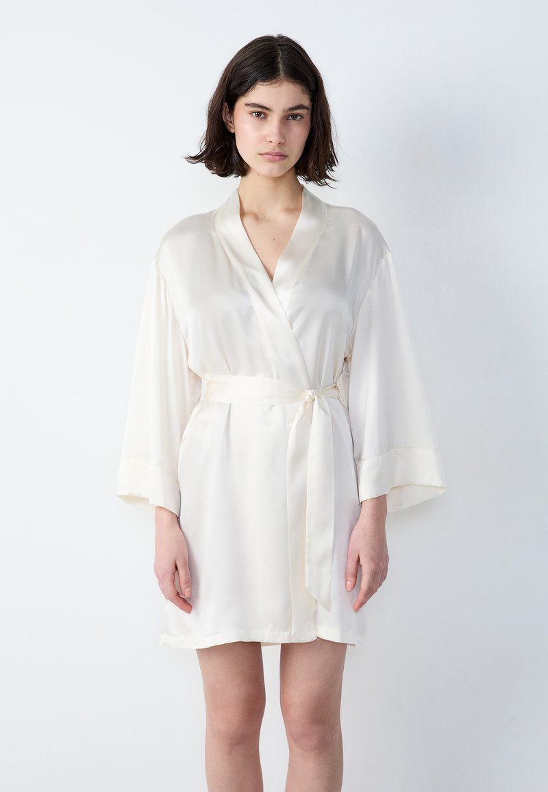 Accappatoio di raso color crema con colletto a scialle, maniche ampie e cintura in vita legata. Tessuto liscio con dettagli di design minimi.