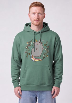 Homme portant un sweat à capuche vert avec un chat licorne de dessin animé entouré de branches feuillues et de petits cœurs, debout devant un fond uni.
