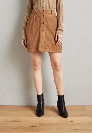Brązowa spódnica sztruksowa z dopasowaną talią, guzikami z przodu i bocznymi kieszeniami, w połączeniu z czarnymi botkami na kostkę na drewnianej podłodze.