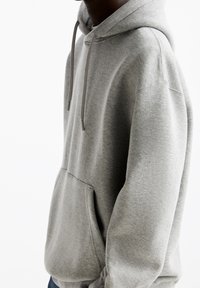 Sweat à capuche gris avec une poche kangourou, des cordons de serrage, une texture en molleton doux et des poignets côtelés. Présente une coupe simple et détendue.