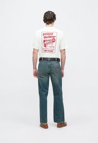 Λευκό κοντομάνικο μπλουζάκι με κόκκινο γραφικό σχέδιο στην πλάτη, σε συνδυασμό με μπλε τζιν τζιν και καφέ παπούτσια. Ίσια γραμμή με τσέπες.